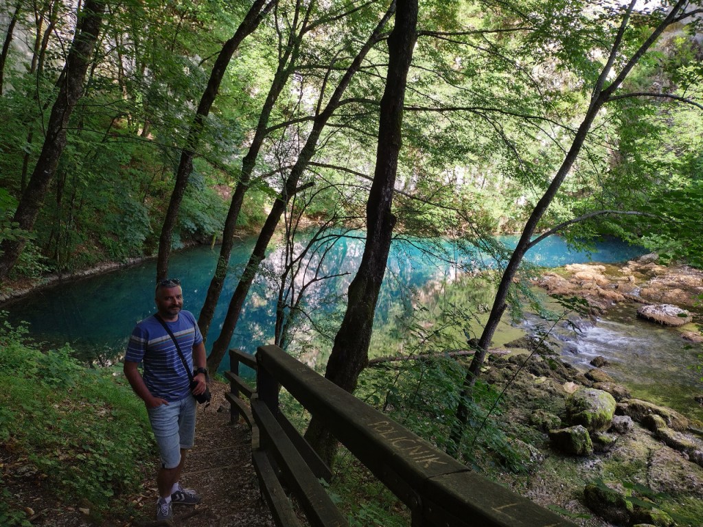 A forty-something man wearing a blue T-shirt and pale blue shorts is standing by a small woodland pond surrounded by thick, green undergrowth, bushes and spindly trees. The turquoise water of the pond slowly flows into a stream to the right. In the background, a hint of steep karst rocks. 