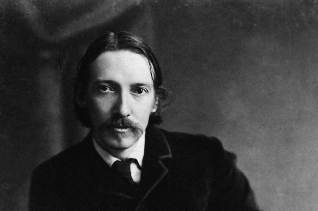 Black-and-white portrait of a man with medium-length hair parted in the middle and a mustache, wearing a dark suit jacket, white shirt, and tie. The plain background and soft lighting emphasize his face and upper body, giving the image a vintage, late 19th–early 20th century style.