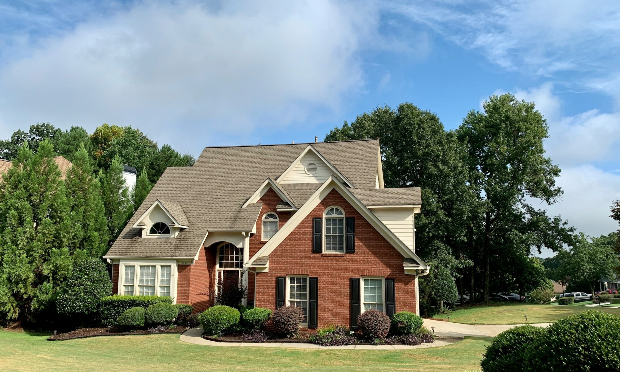 Two‑story suburban house with red brick and beige siding exterior, steep brown shingle roof, multiple gables, arched windows with black shutters, and a landscaped front yard featuring trimmed bushes, garden beds, and tall trees under a partly cloudy sky.