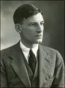 Black-and-white studio portrait of a person in a formal suit with jacket, vest, collared shirt, and tie, short neatly combed hair, facing slightly to the side against a softly blurred background.